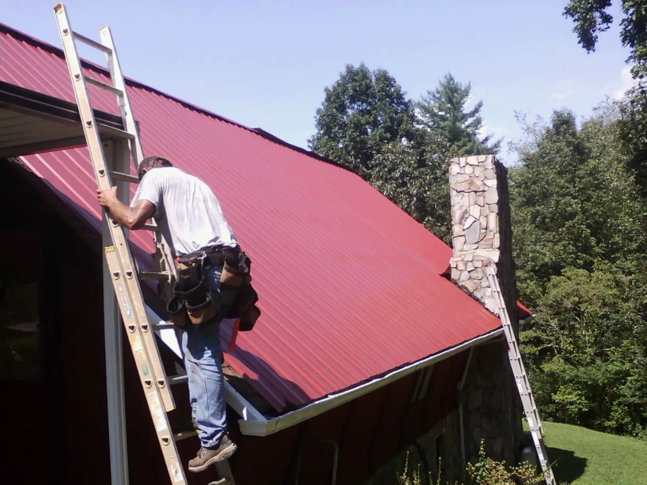 Roofer on a ladder beside a red metal roof and stone chimney — Soffit & Fascia Repair by Roofing Brothers in Waterville